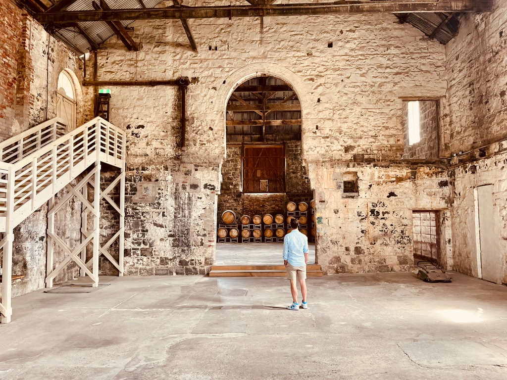 Cam Hamilton stands in a large, rustic, and historic industrial space with exposed stone walls, wooden beams, and a high ceiling. A white staircase with a wooden railing ascends to a mezzanine on the left. In the center, an arched doorway reveals a row of wooden barrels in the background, adding depth to the scene. Light streams in through small windows, highlighting the textured surfaces and raw character of the space. The person, wearing a light blue shirt, beige shorts, and blue shoes, appears to be observing the architecture.