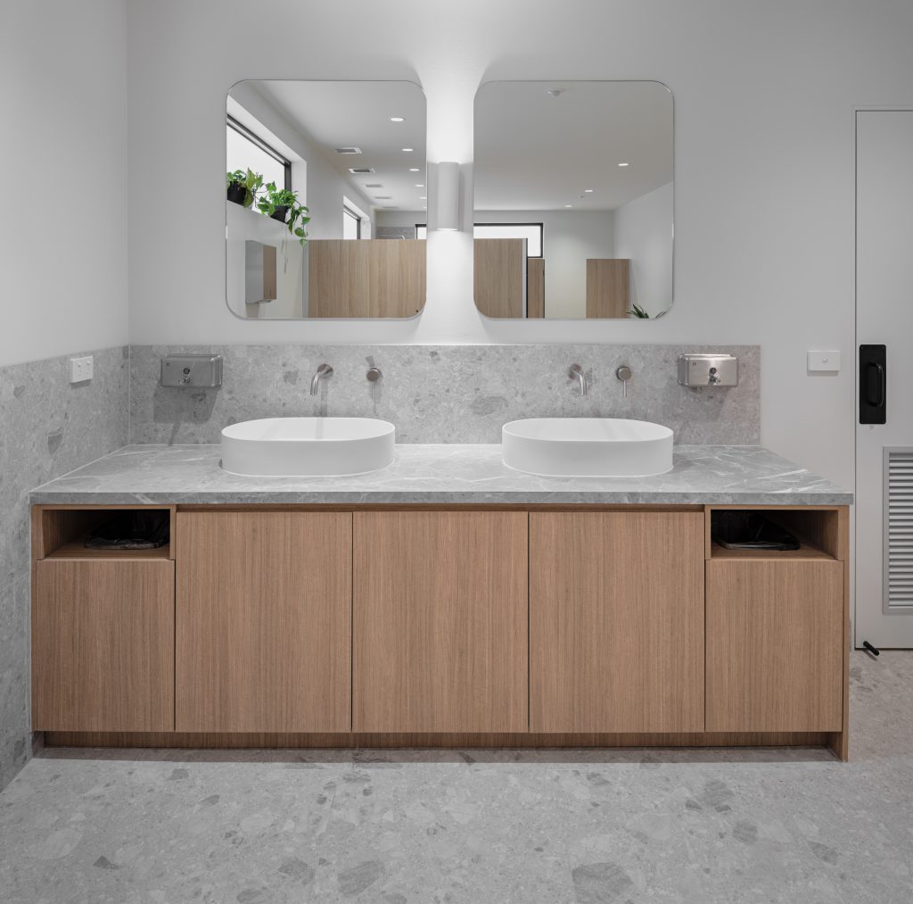 Office fit-out in Laverton North by Hamilton Group. Modern bathroom featuring light timber cabinetry, a white countertop basin with sleek silver tapware, and grey marble-look tiles on both the splashback and floor, creating a clean and contemporary aesthetic.